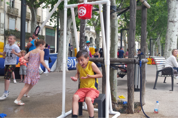 un enfant assis sur un tabouret avec au-dessus un seau d'eau qui lui tombe dessus.